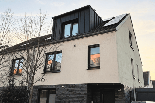 Moderne Wohnhausfassade mit großen Fenstern und mit einer modernen Gaube. Bäume im Vordergrund. Wolkiger Himmel.